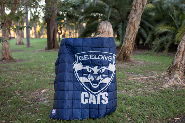 Navy quilted puffy blanket featuring Geelong Cats logo and text, draped over person's shoulders outdoors