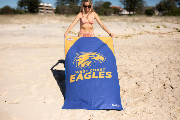 Blue West Coast Eagles beach towel with yellow eagle logo, held by person on sand at beach