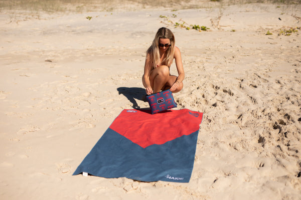 Red and navy colorblock beach towel with AFL branding, displayed on sand with matching patterned pouch.