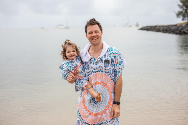 Man and toddler wearing matching blue and coral patterned hooded towels at the beach.