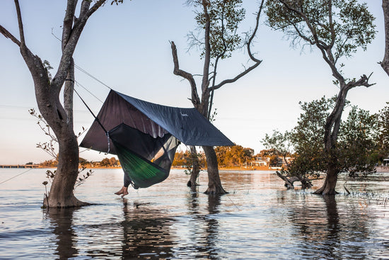 Person relaxing in a Nakie hammock, bug net, and tarp set up between trees over shallow water at sunset.