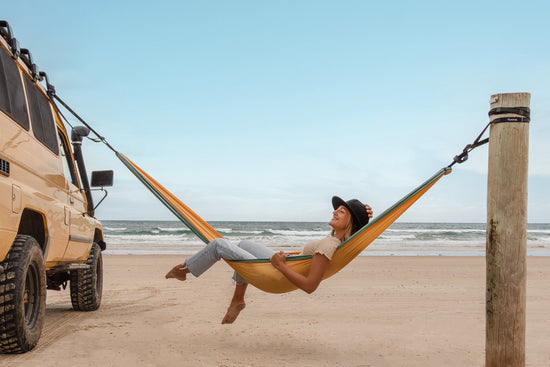 Woman relaxing in a Nakie hammock on the beach, strung between a 4WD vehicle and a wooden post with the ocean in the background.