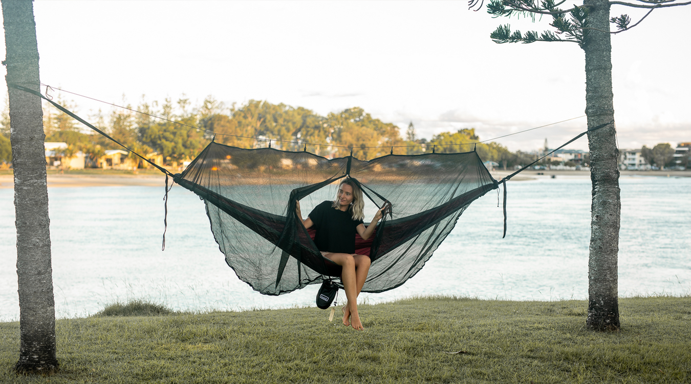 Woman in a Nakie hammock with a bugnet relaxing by the water.