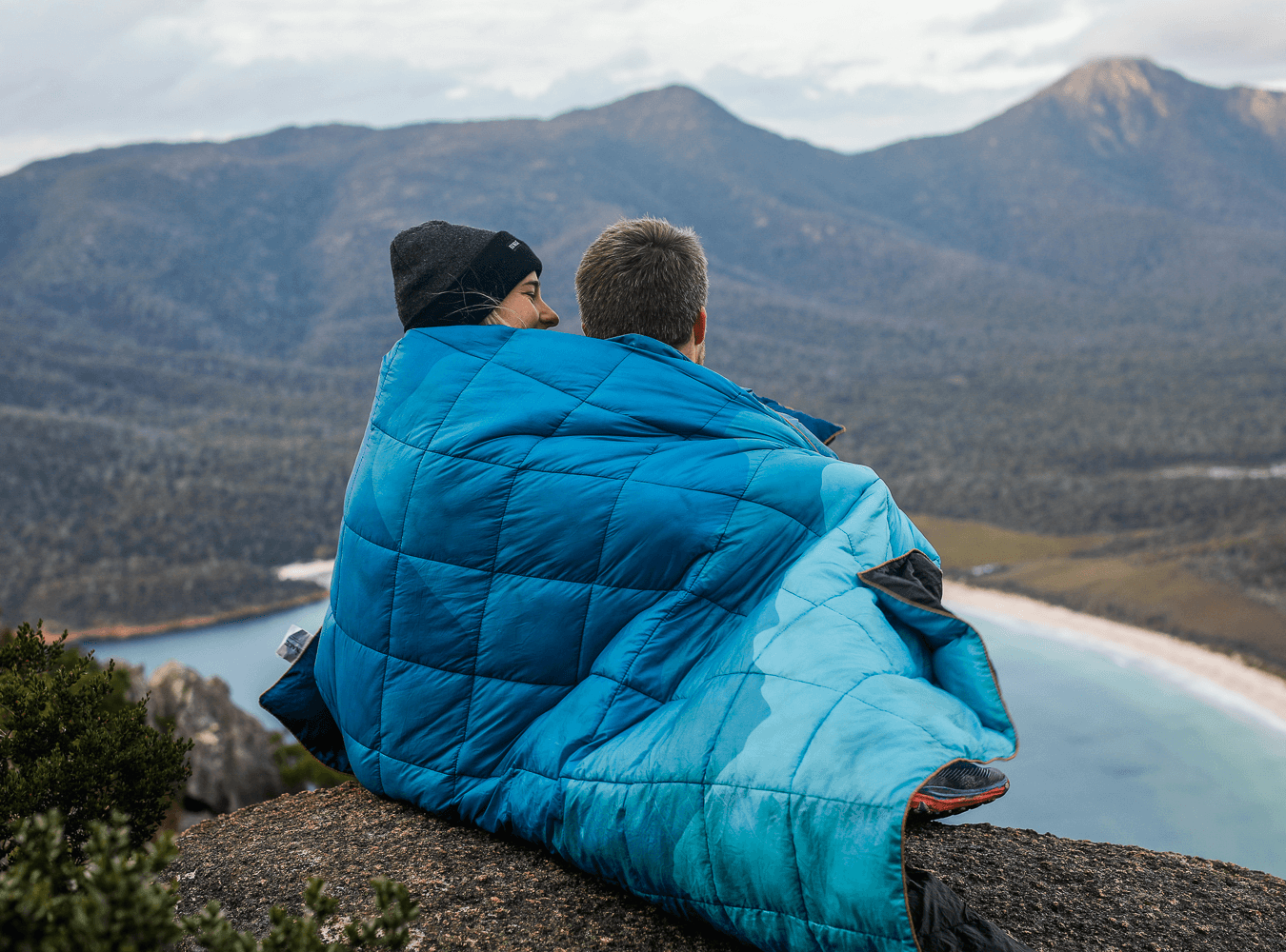 Two people wrapped in a blue puffy blanket sitting on a mountain edge