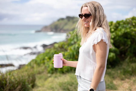 Dusty pink insulated coffee cup lid held by woman in white shirt at beach outlook