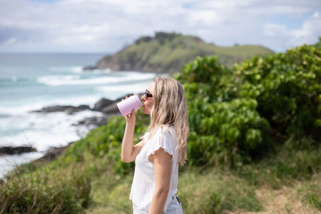 Dusty pink coffee cup lid shown being held by woman drinking outdoors with ocean cliff backdrop