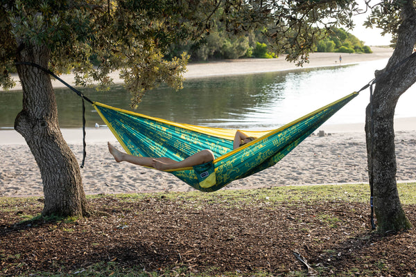 Teal and yellow patterned camping hammock suspended between two trees with black straps, person relaxing by river.