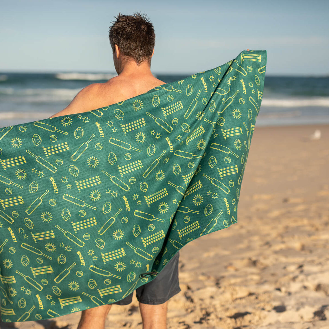 Green and yellow cricket-themed recycled beach towel with sun and bat pattern, shown draped on person at beach.
