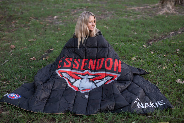 Black quilted puffy blanket with Essendon Bombers red and white graphic design, shown in use outdoors on grass.