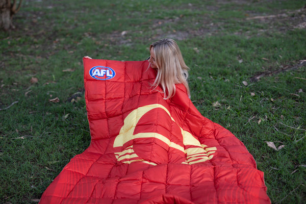 Red AFL Gold Coast Suns puffy blanket with gold curved stripes and logo, laid on grass with woman holding corner.
