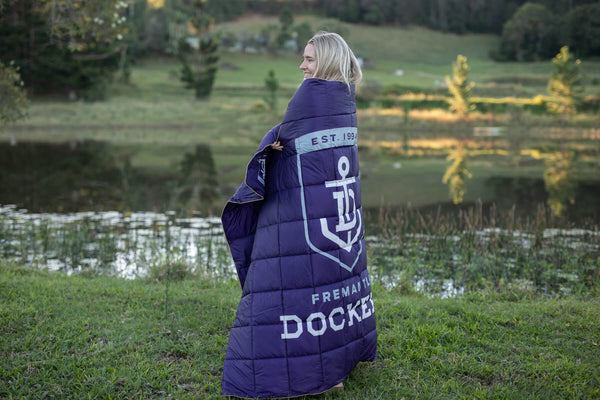 Navy puffy down blanket with Fremantle Dockers logo and "EST. 1994" text, shown draped over person's shoulders outdoors.