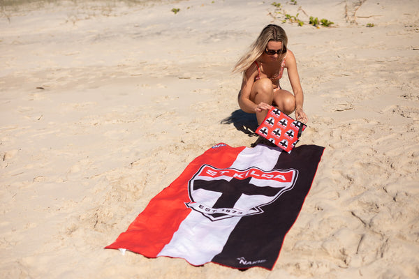 St Kilda Saints AFL beach towel in red, white and black with emblem, displayed on sand with woman holding matching pillow.