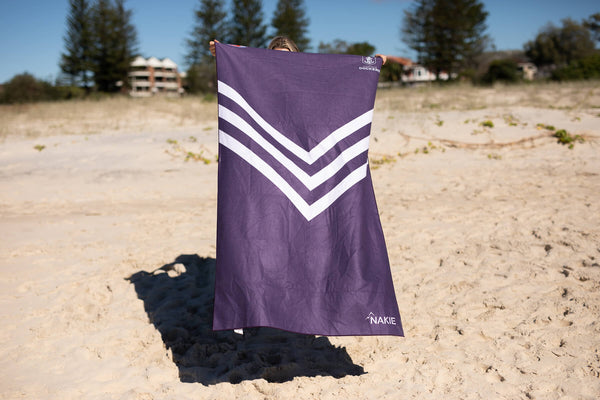 Purple beach towel with white chevron stripe design, held up on sand with beach in background