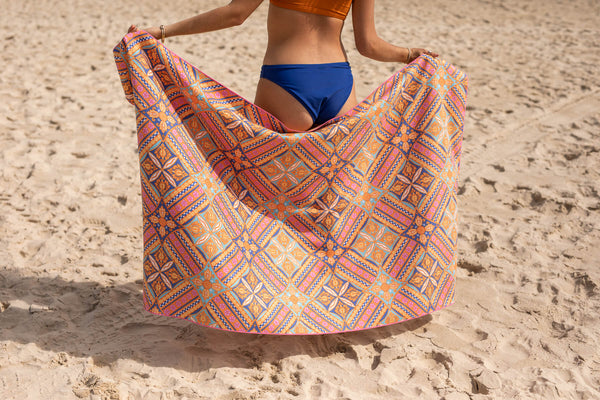 Multicolored geometric patterned beach towel in orange, pink, and blue tones, displayed on sand.