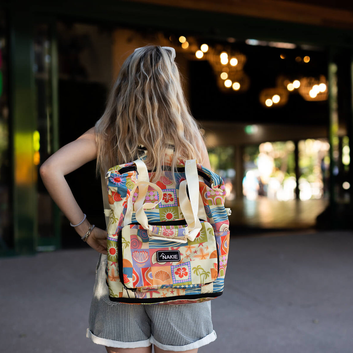 Colorful retro-print recycled tote bag with cream canvas handles, worn on back by person in urban setting.