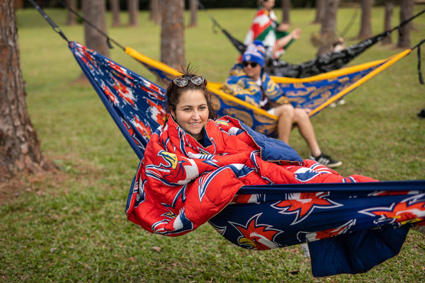 Red and navy rooster-print recycled hammock with integrated straps, shown in use between trees.