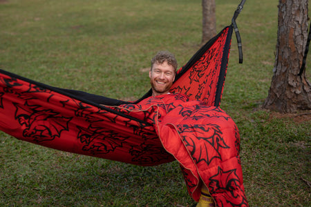 Man smiling in a red NRL Dragons Nakie hammock with black patterns on grass
