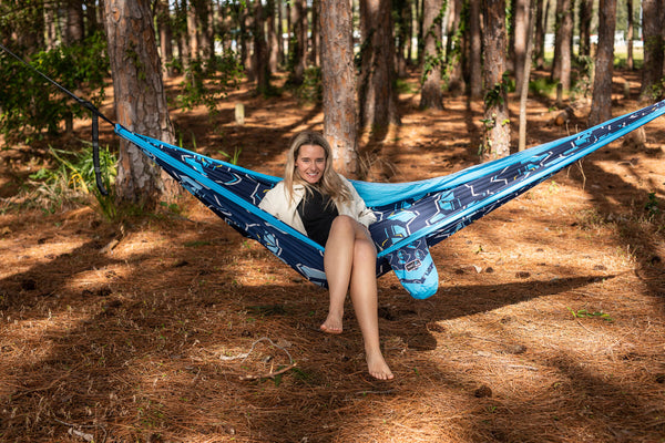 Blue and navy patterned recycled hammock with straps, suspended between forest trees with woman relaxing inside.