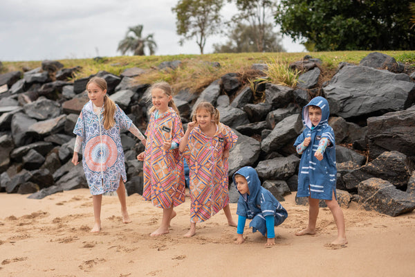 Kids wearing patterned hooded towels and colorful beach cover-ups on sandy beach with rocks behind them
