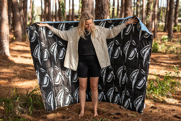 Black and white leaf-patterned puffy blanket with blue trim, displayed open outdoors by model in forest setting