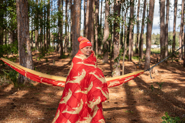 Red puffy blanket with cream dolphin print, worn over shoulders in forest setting with matching red and gold beanie