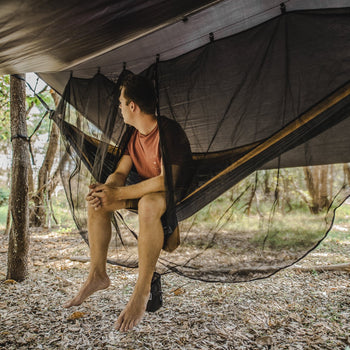 A man sitting comfortably in a Nakie hammock, protected by our Bug Net & Rain Tarp Combo, showcasing the ultimate outdoor relaxation experience with complete protection from insects and rain.