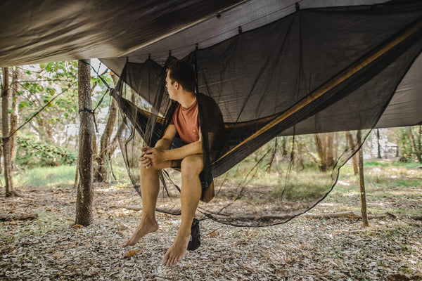 A man sitting comfortably in a Nakie hammock, protected by our Bug Net & Rain Tarp Combo