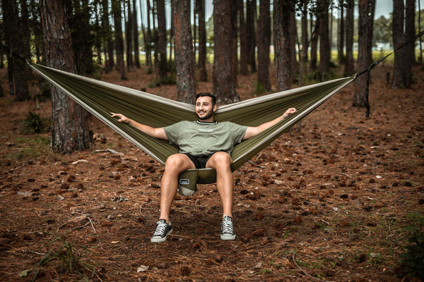 Olive-green camping hammock with integrated pillow suspended between two forest trees, occupied by smiling man